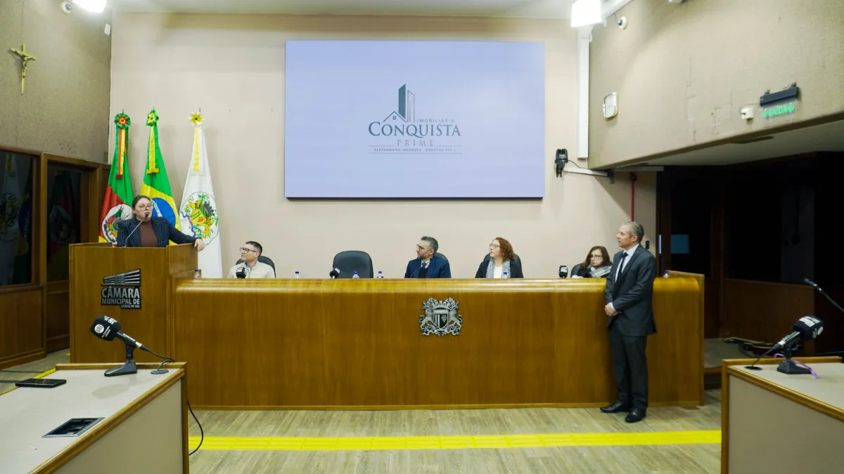 Mesa da sessão solene com a marca da Imobiliária Conquista projetada no telão do plenário
